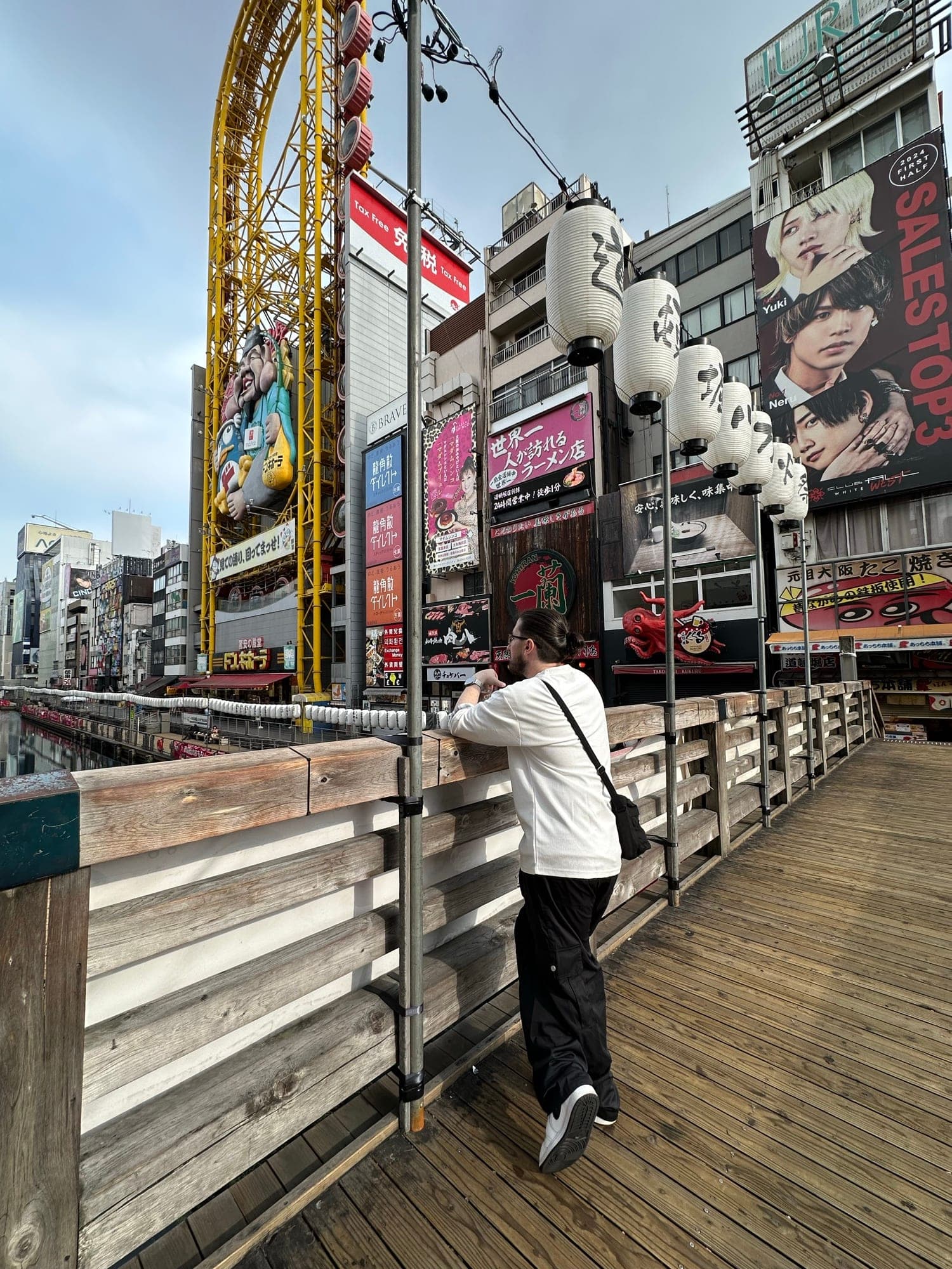 Dave leaning on a wooden bridge railing, looking out at the neon signs and giant Ferris wheel of Dotonbori